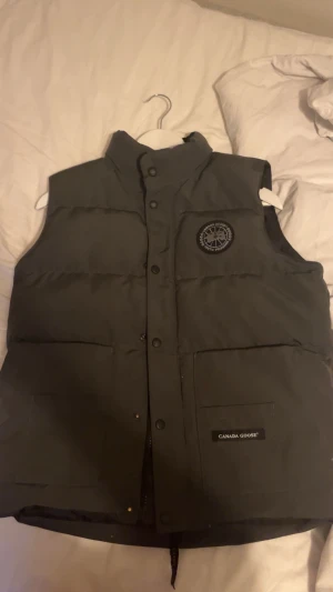 Svart dunväst från Canada Goose - Säljer en svart dunväst från Canada Goose med hög krage och snyggt broderat märke på bröstet. Västen har två stora fickor framtill, knappar och dragkedja. Perfekt för kalla dagar när du vill hålla stilen. Den är äkta!! Ny pris 6999kr, behöver få sålt den snabbt så mitt pris e endast 3699kr