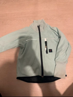 Ljusgrön fleecejacka Polarn O. Pyret - Mysig ljusgrön fleecejacka från Polarn O. Pyret med svart dragkedja och hög ribbad krage. Jackan har en bröstficka med dragkedja och en liten logotyp på bröstet. Perfekt för lager-på-lager under kyliga dagar. Storlek 98
