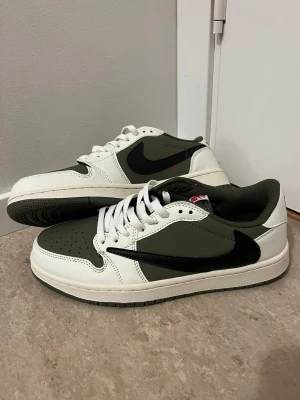 Nike Air Jordan 1 Low Travis Scott Olive - Nike Air Jordan 1 Low Travis Scott i olivgrönt, vitt och svart skinn. Swoosh-loggan är svart och vänd åt fel håll, med röda broderade detaljer på hälen. Klassisk snörning och perforerad tå. Snygg och unik sneaker med Travis Scott smiley på baksidan.