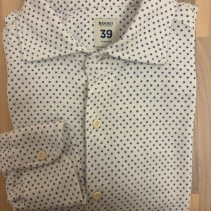Vit skjorta med blå prickar från Boggi - Snygg vit skjorta från Boggi Milano med blått prickigt mönster. Tillverkad i mjuk och len bomull som känns skön mot huden. Klassisk krage, långa ärmar och knäppning framtill. En tidlös skjorta som fungerar lika bra till jeans som till chinos. Enbart testad.  Mått:	Armhåla till armhåla: 46 cm,	Axel till axel: 54 cm, Topp till botten: 48 cm,	Axel till ärmslut (cuff): 64 cm