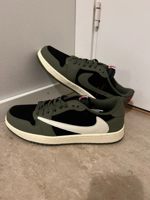 Nike Air Jordan 1 Low Travis Scott Olive - Nike Air Jordan 1 Low Travis Scott i olivgrönt och svart med vit omvänd swoosh. Skorna har röda broderade detaljer på hälen och klassisk snörning. Ovandelen är i skinn och mocka, med en platt sula i beige. Perfekt för dig som gillar streetwear och unika sneakers.