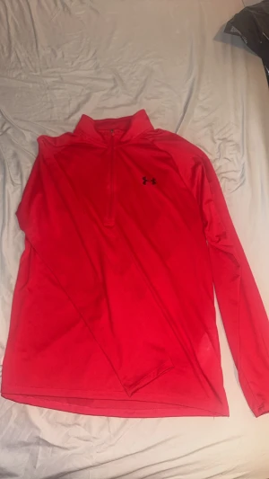 Röd träningströja från Under Armour - Snygg röd långärmad träningströja från Under Armour med halv dragkedja och svart logga på bröstet. Tröjan har en loose fit och är tillverkad i ett mjukt, syntetiskt material som andas. Perfekt för träning eller sportiga dagar. Helt ny använd 1 gång, storlek L passar M