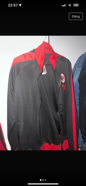 AC Milan x Puma svart och röd jacka - AC Milan track jacket från Puma i svart och rött med klubbmärke på bröstet och Puma-logga. Jackan har dragkedja framtill, ståkrage och röda detaljer längs ärmar och axlar. Perfekt för dig som älskar fotboll och vill visa ditt lagstöd.