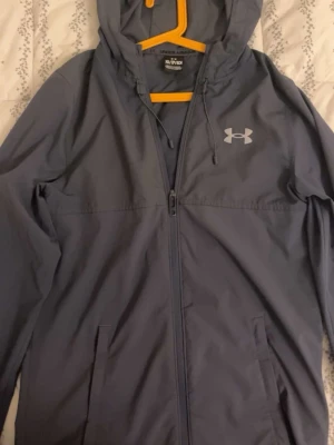 Blå vindjacka från Under Armour XS - Snygg blå vindjacka från Under Armour i storlek XS. Jackan har huva med snörning, dragkedja framtill och två fickor. Stort Under Armour-tryck på ryggen och logga på bröstet. Perfekt för aktiva dagar och sportiga outfits. Pris kan diskuteras.