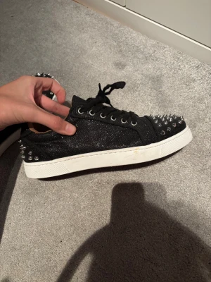 Louboutin skor - Säljer ett par svarta sneakers från Louboutin med glittrigt tyg och silverfärgade nitar på tå och häl. Skorna har snörning, vit platt sula och den klassiska röda undersidan. Riktigt edgy och sticker ut med sin coola design.