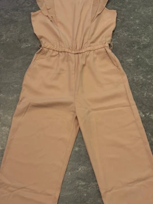 Beige byxdress med volangdetalj - Snygg beige byxdress med breda ben och volangdetaljer vid axlarna. Resår i midjan för skön passform och praktiska fickor på sidorna. Perfekt för dig som gillar en avslappnad men trendig look.