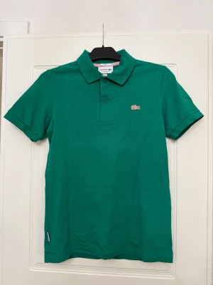 Grön piké från Lacoste - Säljer en klassisk grön pikétröja från Lacoste med korta ärmar och krage. Tröjan har två knappar framtill och den ikoniska krokodilloggan broderad på bröstet. Tillverkad i mjuk bomull som är skön mot huden. Perfekt för dig som gillar stilren och sportig look.
