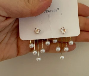 Örhängen med pärlor och strass W Jewelry - Snygga örhängen från W Jewelry med glittrande strass i mitten och hängande vita pärlor på tunna guldfärgade kedjor. Designen är modern och unik, perfekt för dig som vill sticka ut. Kommer med extra pärlpluppar för variation.