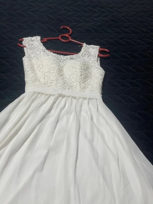 Vit lång spetsklänning med v-ringning - Elegant långklänning i vitt med spetsliv och v-ringad rygg. Överdelen har ett detaljerat blommigt spetsmönster och kjolen är i ett mjukt, flytande tyg. Klänningen är ärmlös och har ett markerat midjeband för en snygg siluett. den är bara använd en gång.