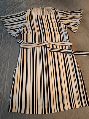 Randig kortklänning från H&M - Snygg kortklänning från H&M med svartvita vertikala ränder och volangärmar. Klänningen har rund halsringning och ett matchande bälte i midjan för extra stilpoäng. Perfekt för dig som gillar en fräsch och modern look.