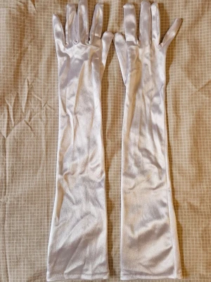 Långa vita satinhandskar - Långa handskar i vit satin. Handskarna är släta och glansiga med en mjuk känsla och går upp över armbågen för en classy look. Perfekta för att lyfta din outfit till nästa nivå.
