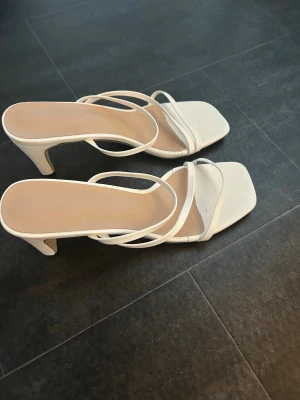 Vita sandaler med klack och smala remmar - Säljer ett par stilrena vita sandaler med smala remmar och fyrkantig tå. Skorna har en elegant hög klack och öppen tå, vilket ger en modern vibe. Perfekta för dig som gillar minimalistisk design och vill ha något som sticker ut. Det är storlek 41, men passar mig som vanligtvis har 38/39. 