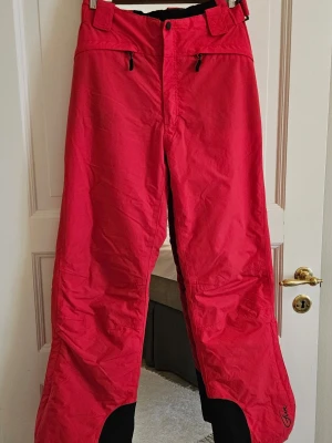 Röda täckbyxor från Five med hängslen - Riktigt snygga röda täckbyxor från Five med svarta förstärkningar vid bensluten och justerbara svarta och vita hängslen med logga. Byxorna har dragkedjor vid fickorna och bred passform, perfekta för vinterns äventyr.