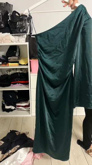 Mörkgrön långklänning med oneshoulder - Säljer en elegant mörkgrön långklänning i glansigt material med oneshoulder och lång ärm. Klänningen har en snygg drapering och faller fint längs kroppen, perfekt för dig som vill sticka ut med en unik siluett.