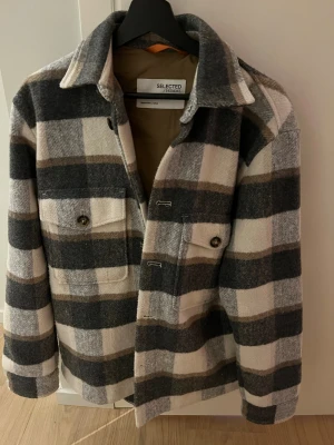 Rutig jacka från Selected Homme - Snygg overshirt från Selected Homme i rutigt mönster med grå, beige och vita toner. Jackan har två bröstfickor med knappar, klassisk krage och stängs med knappar framtill. Perfekt lager-på-lager-plagg för höst och vår. Den passar perfekt till både höst och vinter