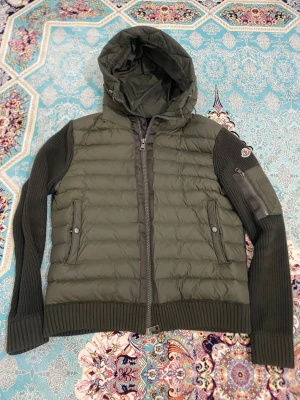 Moncler cardigan - Snygg olivgrön pufferjacka från Moncler med stickade ärmar och quiltad front. Jackan har huva, dragkedja framtill, fickor med tryckknapp och Moncler-logga på ärmen. Perfekt för kyliga dagar. Obs det är fastpris.