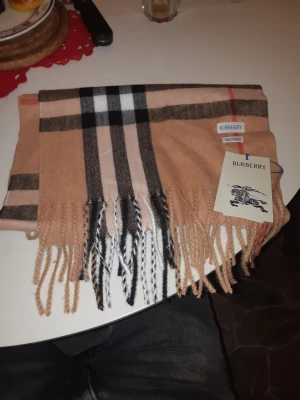 Rutig beige halsduk från Burberry - Snygg väst från Burberry i klassiskt rutigt mönster med beige, svart, vitt och röda detaljer. Västen har fransar längs nederkanten och är tillverkad i mjukt material som ger en lyxig känsla. Perfekt för dig som gillar ikonisk design och vill sticka ut.