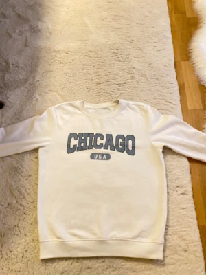 Vit sweatshirt med Chicago-tryck - Vit sweatshirt med stort blått tryck 'CHICAGO USA' på bröstet. Tröjan har rund hals, ribbade muddar och lång ärm. Perfekt för dig som gillar college-vibe och enkel stil. Materialet känns som mjuk bomull och passformen är normal. Helt ny har haft på mig den 5 gånger sen des har den bara legats i garderoben Hör av er om ni har frågor pris kan diskutera 