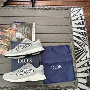 Grå Dior sneakers med reflekterande detaljer - Säljer ett par grå Dior sneakers med stora reflekterande CD-loggor på sidorna. Skorna har vit och svart sula, mesh- och syntetpaneler samt rund tå. Kommer med originalkartong och dustbag. Perfekta för dig som vill sticka ut med exklusiva sneakers.