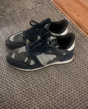 Säljer ett par Valentino Rockrunner sneakers i mörkblått och beige med snyggt camouflage-mönster. Skorna har snörning, platt sula och detaljer i mocka och mesh. Ikoniska nitar på hälen och rund tå ger en cool vibe. Perfekta för dig som gillar streetstyle och designer.