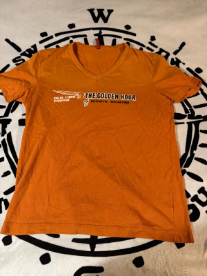 Nudie Jeans t-shirt orange med ”THE GOLGDEM HOUR”  tryck   - Snygg orange t-shirt från Nudie Jeans med v-ringning och  tryck på bröstet: 'The Golden Hour'. Tillverkad i 100% bomull, kortärmad och med en normal passform. Perfekt för dig som gillar streetwear och vill sticka ut med färg.