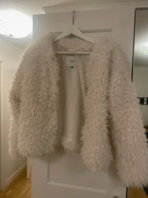 Vit pälsjacka från Jacqueline de Yong - Fluffig och trendig vit pälsjacka från Jacqueline de Yong. Jackan har lång ärm, öppen front och är tillverkad i ett mjukt syntetmaterial som ger en cool och mysig look. Perfekt för dig som vill sticka ut med en statementjacka.