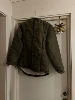 Olivgrön pufferjacka med fuskpälsfoder - Olivgrön pufferjacka med huva och vit, mjuk fuskpäls på insidan. Jackan har knappar och dragkedja framtill, två fickor med lock och elastisk midja bak för snygg passform. Perfekt för kalla dagar när du vill vara både varm och stilren. Aldrig använd, bara testad. Mvh Liam 