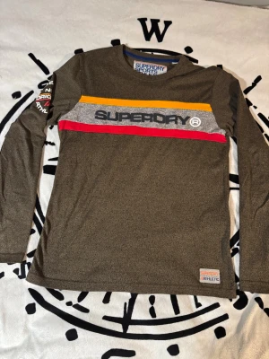 Superdry långärmad tröja med tryck -  långärmad tröja från Superdry i brun färg med breda ränder i grått, gult och rött över bröstet. Stor Superdry-logga framtill och patch nertill. Rund hals och tryck på ena ärmen. Perfekt för dig som gillar sportig streetstyle.