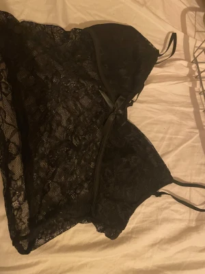 Svart spets linne med rosett - Säljer en svart linne med tunna axelband och vacker spets över hela toppen. Den har en liten rosett framtill som detalj och är genombruten med ett feminint mönster. Perfekt för dig som gillar en edgy och stilren look.