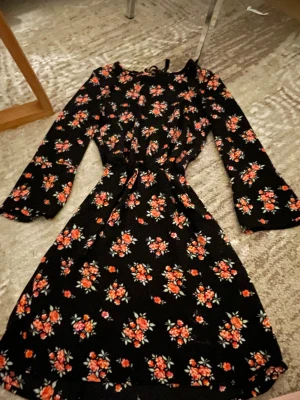 Blommig svart klänning H&M Divided - Supersöt svart kortklänning från H&M Divided med blommigt mönster i rött och rosa. Klänningen har långa ärmar, knytning i nacken och markerad midja. Perfekt för dig som gillar romantiska vibes och vill sticka ut lite extra.