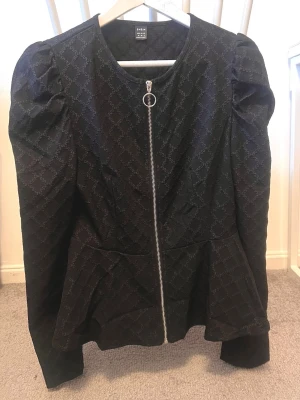 Svart mönstrad jacka från SHEIN - Snygg svart jacka från SHEIN med diskret rutmönster och rund hals. Jackan har puffade axlar och en silverfärgad dragkedja framtill med ringdetalj. Passformen är normal och materialet känns som polyester.