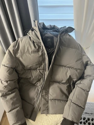 Grå pufferjacka från Canada Goose - Säljer en grå pufferjacka från Canada Goose med huva och snyggt broderat märke på ärmen. Jackan har dragkedja framtill, ribbade muddar och är fodrad med mjukt material. Perfekt för kalla dagar och har en clean, modern look. Storlek S men passar som M. Säljer den då den va lite för stor för mig och endast testad