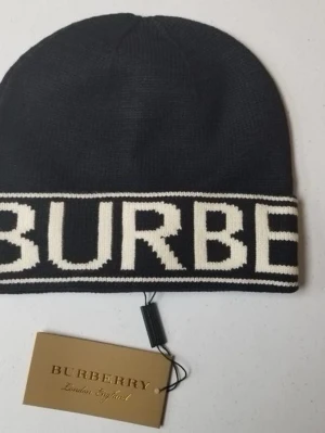 Svart Burberry mössa med logga - Snygg svart mössa från Burberry med stor vit logga längs kanten. Mössan är stickad och har en klassisk form som passar perfekt för kyliga dagar. Stilren design med kontrastfärg på loggan gör den till ett statement-plagg.