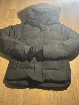 Svart pufferjacka från Canada Goose - Svart pufferjacka från Canada Goose med huva och snygg logotyp på ärmen. Jackan har dragkedja framtill och två praktiska fickor. Materialet är polyester och bomull, med dunfyllning för extra värme. Perfekt för kalla dagar och riktigt stilren look.