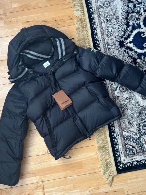 Svart pufferjacka från Burberry - Svart pufferjacka från Burberry med avtagbara ärmar, vilket gör att du kan bära den som väst. Jackan har en stor huva med grått och vitt randigt foder, dragkedja och knappar framtill samt praktiska fickor med dragkedja. Perfekt för kalla dagar och riktigt snygg streetstil.