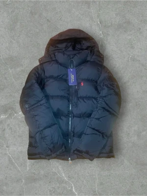 Svart pufferjacka från Polo Ralph Lauren - Svart pufferjacka från Polo Ralph Lauren med huva och dragkedja framtill. Jackan har en klassisk puffer-design med quiltade sektioner och en liten röd logga på bröstet. Perfekt för kalla dagar och har en snygg, enkel look.