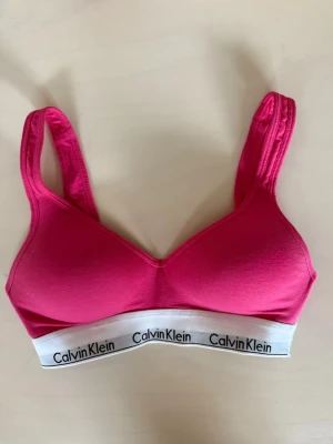 Rosa Calvin Klein sport-bh - Sportig rosa bh från Calvin Klein med bred vit resår och svart logga runt om. Mjuk och stretchig modell med vadderade kupor och breda axelband för extra komfort. Perfekt för en aktiv vardag eller träning.