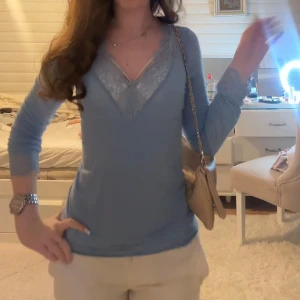 Blå långärmad topp med spets - Snygg blå långärmad topp med v-ringning och spetsdetaljer runt halsen och ärmsluten. Toppens material känns mjukt och stretchigt, perfekt för att skapa en chill och feminin look. Passar dig som gillar stilrena och romantiska detaljer.