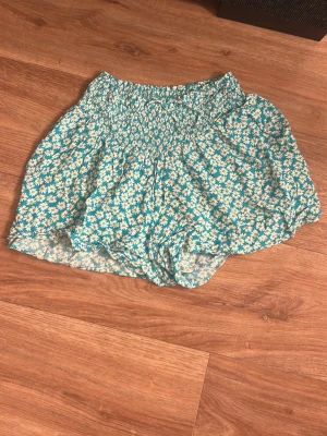 Blommiga turkosa shorts/kjol  - Säljer ett par turkosa shorts med vita och gröna blommor, bred smock i midjan och luftig passform. Perfekta för varma dagar och riktigt sköna att ha på sig. Materialet är mjukt och känns som bomull. Supersöta och somriga!