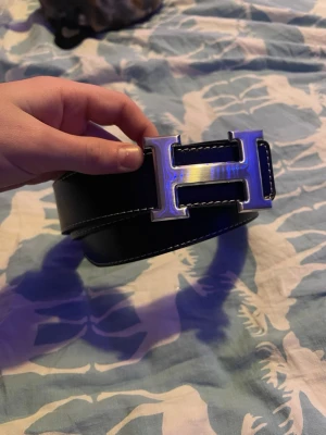 Svart skinnbälte med H-spänne hermess - Snyggt svart bälte i skinn med ett stort, silvrigt H-format metallspänne. Bältet har vit kontrastsöm längs kanterna och är tillverkat i ett grovt, strukturerat läder. Perfekt för att ge outfiten en extra touch. Passar midjemått ca 105 cm.