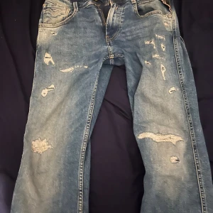 Replay Anbass med slitningar - Storlek 28/32 men sitter som 30/32. Säljer ett par Replay Anbass slim fit jeans i ljusblå tvätt med slitna detaljer och patchade hål på benen. Klassisk femficksmodell med dragkedja och Replay-läderpatch bak i midjan. Perfekt för dig som gillar en edgy streetstil.
