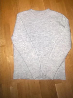 Ljusgrå stickad tröja - En enkel och stilren ljusgrå stickad tröja med rund hals och långa ärmar. Perfekt för att hålla sig varm under kyliga dagar. Tröjan har ribbade muddar vid ärmslut och nederkant för extra komfort.