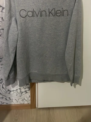 Grå sweatshirt från Calvin Klein - Säljer en stilren grå sweatshirt från Calvin Klein i storlek S. Tröjan har en klassisk passform med rund halsringning och långa ärmar. Stor Calvin Klein-logga framtill ger en clean look. Perfekt till jeans eller joggers.