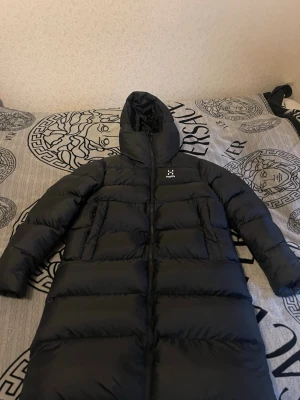 Svart lång dunjacka från Haglöfs - Säljer en svart lång dunjacka från Haglöfs i storlek M. Jackan har huva, tvåvägsdragkedja och stora fickor. Perfekt för kalla dagar och riktigt skön att bära. Klassisk design med diskret logga på bröstet.