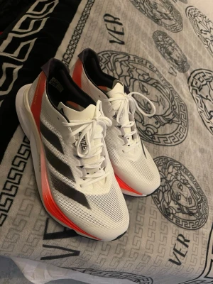 Adidas Adizero Lightstrike Pro sneakers - Säljer ett par Adidas Adizero Lightstrike Pro sneakers i vitt med svarta ränder och orange detaljer. Ovandelen är i mesh för bra ventilation och sulan är tjock och responsiv, perfekt för löpning. Skorna har snörning och en sportig look.