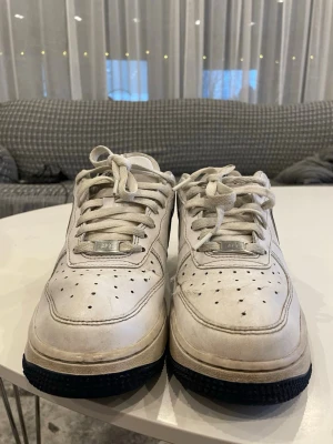 Nike Air Force 1 sneakers vit/blå - Säljer ett par klassiska Nike Air Force 1 sneakers i vitt med mörkblå detaljer och sula. Skorna har snörning, perforerad tå och rund tå. Tillverkade i skinn med syntetfoder. Perfekt för dig som gillar streetstyle och ikoniska sneakers.