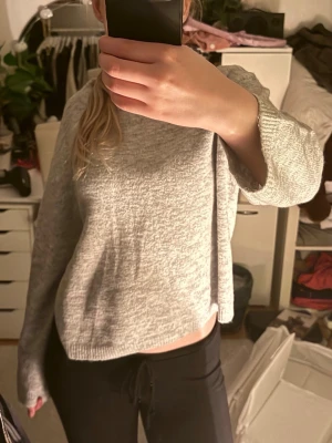 Grå stickad tröja från 157 - Mysig grå stickad tröja från 157 med avslappnad passform och långa ärmar. Tröjan har ribbade muddar vid ärmslut och nederkant, perfekt för lager på lager under kyligare dagar. Enkel och stilren design som funkar till allt.