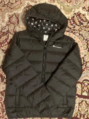 Svart pufferjacka från Champion XL - Svart pufferjacka från Champion i storlek XL. Jackan har huva med logomönster på insidan, dragkedja framtill och två sidofickor. Perfekt för kalla dagar och har en schysst, sportig vibe. Materialet är polyester och jackan är quiltad för extra värme.