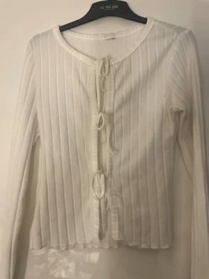 Vit ribbad kofta med knyt - Säljer en söt vit ribbad kofta med långa ärmar och tre knyt framtill. Koftan har en rund halsringning och är gjord i ett mjukt material som känns skönt mot huden. Perfekt att slänga över en topp för en chill look.