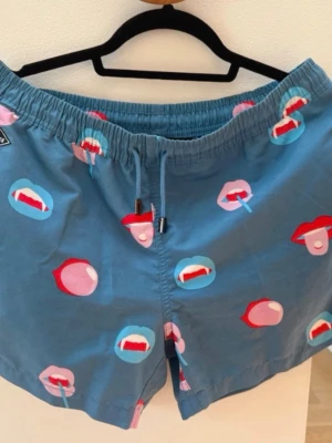 Nikben Blå shorts med läppar och snörning - Säljer ett par blå shorts med elastisk midja och snörning. Shortsen har ett unikt mönster med färgglada läppar i rött, rosa och blått över hela tyget. De är tillverkade i bomull och har en bakficka. Perfekta för sommarens chill dagar.